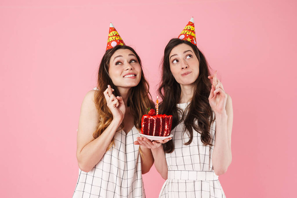 Image of excited women in party cones holding cake and fingers crossed for good luck isolated over pink background - Foto, Imagen