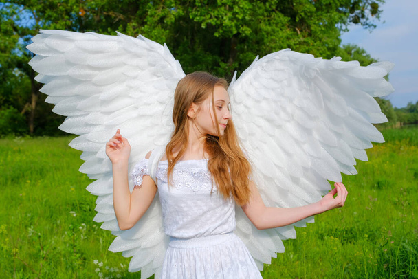 χαριτωμένο καυκάσιος έφηβος κορίτσι με φτερά αγγέλου και λευκό φόρεμα που θέτουν σε εξωτερικούς χώρους στο πάρκο  - Φωτογραφία, εικόνα