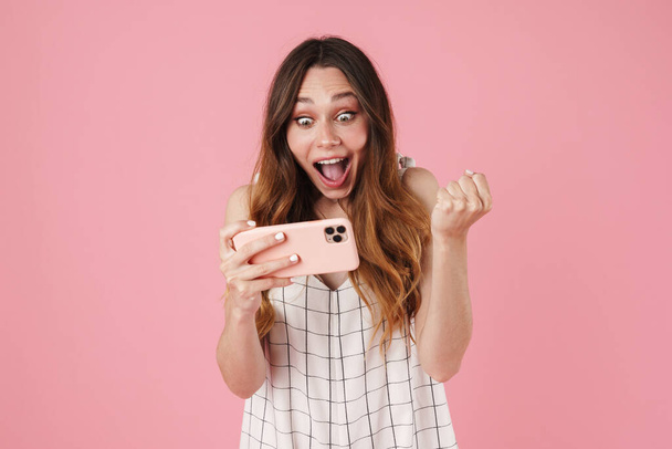 Image of excited cute woman using cellphone and making winner gesture isolated over pink background - Foto, imagen