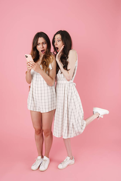 Image of shocked young women screaming and using cellphone isolated over pink background - Φωτογραφία, εικόνα