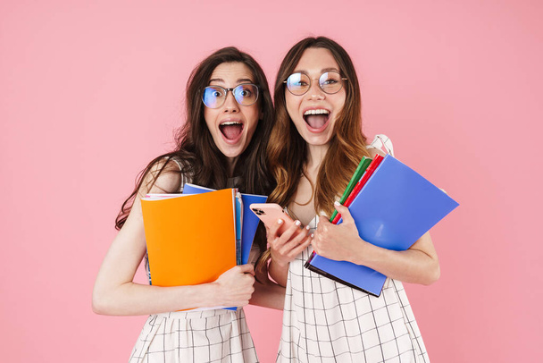 Image of delighted student women holding exercise books and using cellphone isolated over pink background - Photo, Image