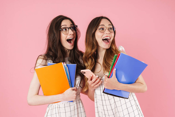 Image of delighted student women holding exercise books and using cellphone isolated over pink background - Fotografie, Obrázek