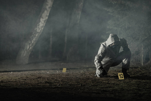 Looking For Traces and Crime Evidence Markers Placing by Forensic Officer in Hazmat Suit and Gloves. Dark Foggy Forest. Crime Scene Theme Concept. - Photo, Image