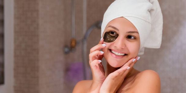 Una mujer sonriente envuelta en una toalla blanca está aplicando parches para los ojos negros después de darse una ducha por la mañana. Quita tus ojeras bajo los ojos
. - Foto, Imagen