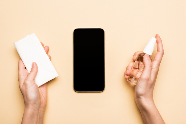 vista parcial de la mujer sosteniendo desinfectante y servilletas de papel cerca de teléfono inteligente con pantalla en blanco en beige
 - Foto, Imagen