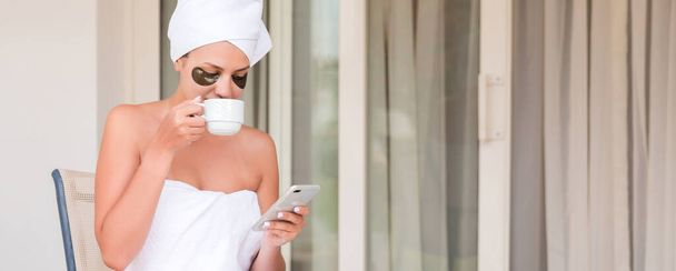 Zabalený do ručníku po sprše žena s páskou přes oko je pití čaje nebo kávy u stolu a čtení zpráv nebo zpráv na hotelové terase. Dívka si užívá dovolenou. - Fotografie, Obrázek