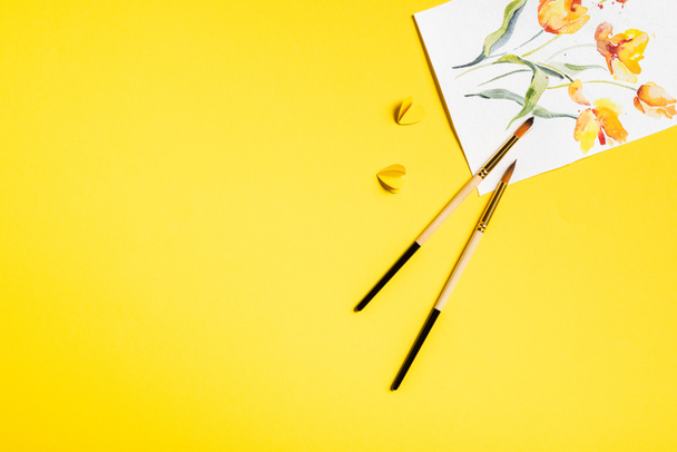 top view of paintbrushes near drawn flowers on painting and paper cut elements on yellow - Foto, afbeelding