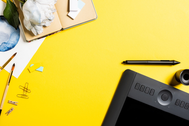 vista superior de la tableta de dibujo con pantalla en blanco, clips, lápiz óptico, pintura, cuaderno y flor en amarillo
  - Foto, Imagen