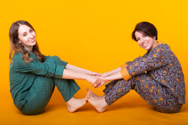 Portret dwóch pięknych młodych dziewcząt ubranych w kolorowe piżamy bawiące się podczas nocowania na żółtym tle. Piżama party i koncepcja wieczoru panieńskiego - Zdjęcie, obraz