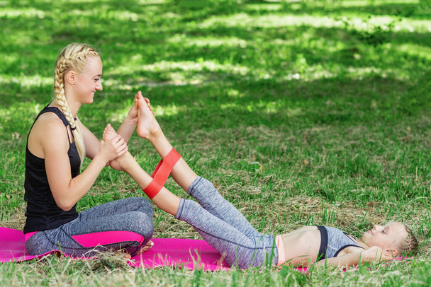 Young woman is training little girl's legs by fitness gum on mat in the park. - Zdjęcie, obraz