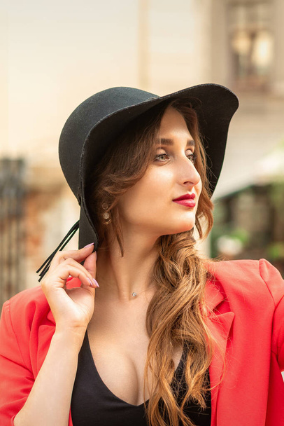 Seitenansicht einer modischen jungen Frau in rotem Mantel und schwarzem Hut, die auf der Straße der Stadt wegschaut. - Foto, Bild