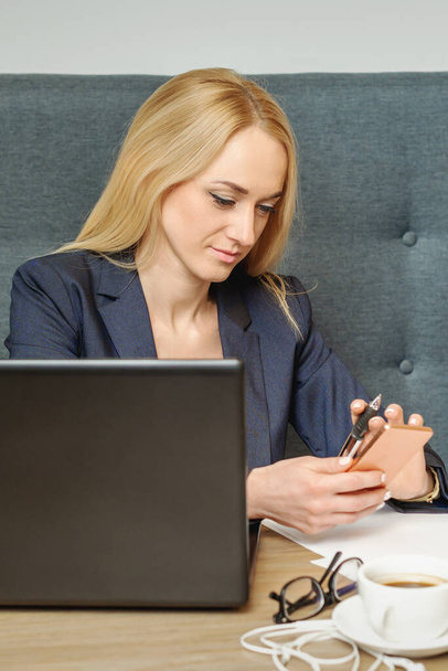 Απασχολημένη ελκυστική γυναίκα με ξανθά μαλλιά χρησιμοποιώντας έξυπνο τηλέφωνο εργασίας με φορητό υπολογιστή στο γραφείο. - Φωτογραφία, εικόνα