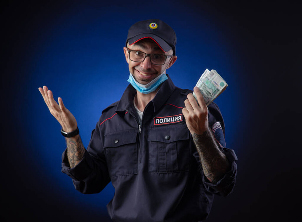Ένας τύπος με στολή αστυνομικού Ρώσος αστυνομικός με ιατρική μάσκα με χρήματα στα χέρια του. Αγγλική μετάφραση του "Police" - Φωτογραφία, εικόνα