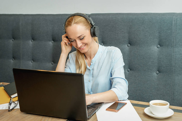 Ευτυχισμένη νεαρή γυναίκα φορώντας ακουστικά κοιτάζοντας στην οθόνη του φορητού υπολογιστή κατά τη διάρκεια της online κατάρτισης με τηλεδιάσκεψη στο γραφείο στο σπίτι. - Φωτογραφία, εικόνα