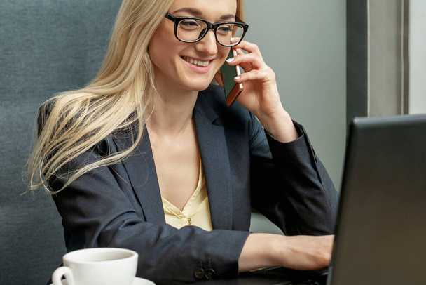 Красивая молодая деловая женщина в очках звонит на мобильный телефон, работает на ноутбуке в домашнем офисе
. - Фото, изображение