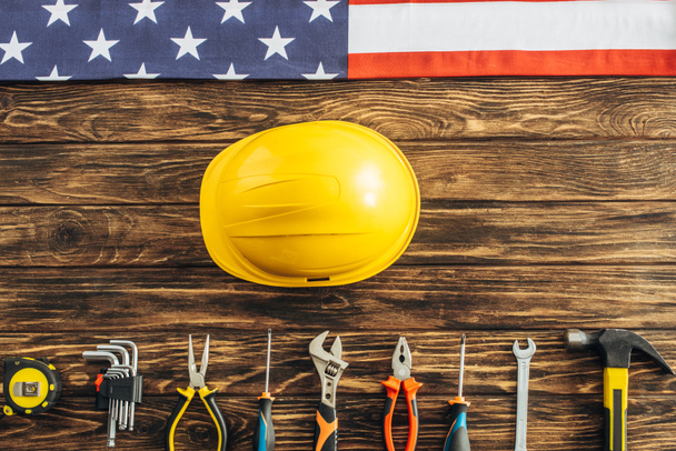 Draufsicht auf metallische Werkzeuge, Schutzhelm und amerikanische Flagge auf Holzoberfläche, Arbeitstagskonzept  - Foto, Bild