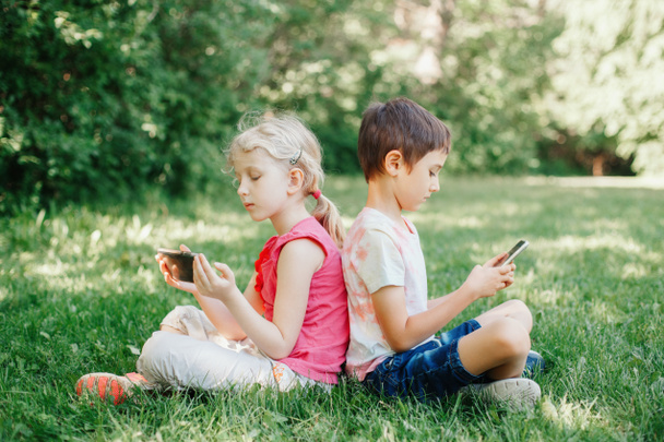 Chlapec a dívka hrát hry na chytrých telefonech venku. Dětské digitální gadget obrazovky závislost. Děti přátelé hrát online sedí na trávě v parku. Současný problém osamělosti společně. - Fotografie, Obrázek