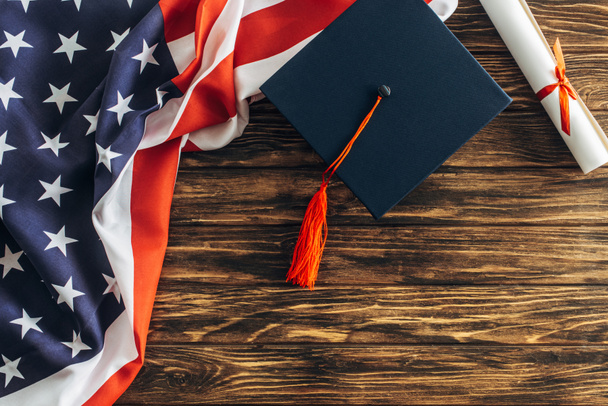 κορυφαία άποψη του διπλώματος και καπέλο αποφοίτησης κοντά αμερικανική σημαία με αστέρια και ρίγες στην ξύλινη επιφάνεια  - Φωτογραφία, εικόνα