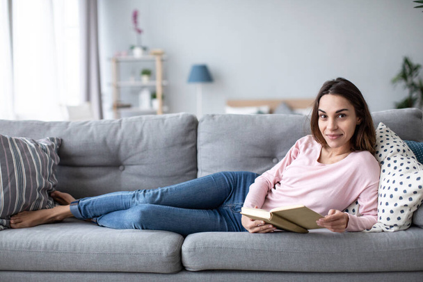 Smiling beautiful woman relaxing on the couch and reading a book in the living room. Alone home. - Foto, Imagem