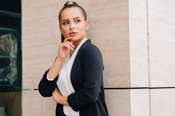 Elegant stylish lady stands in a black austere suit leaning on the wall of the business center. - Photo, Image