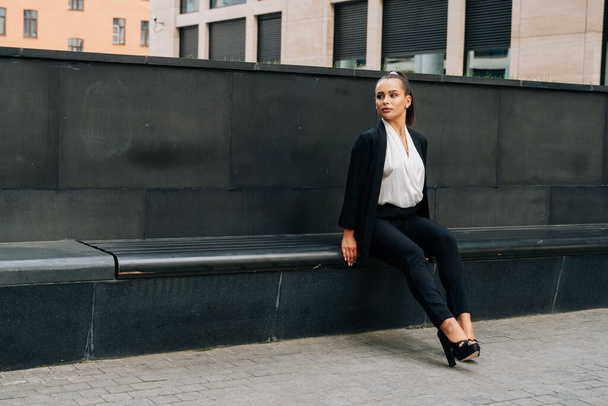 A business lady in a black, stern suit sits on a bench outside work at lunchtime. Customer's waiting concept. - Photo, image