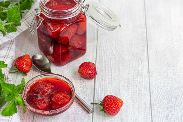 Strawberry jam in a glass jar next to fresh strawberries. On a white wooden background. Homemade winter fruit blanks. Selective focus. Copy space - Photo, Image