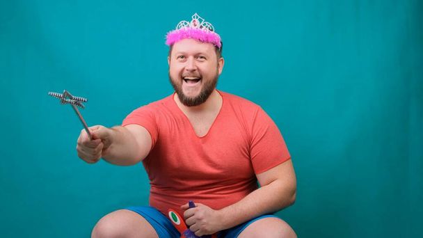handsome bearded freaky man in a pink T-shirt with a diadem on his head is riding astride a unicorn with a magic wand in his hand. A funny wizard joke to make and fulfill a wish. - Foto, Bild