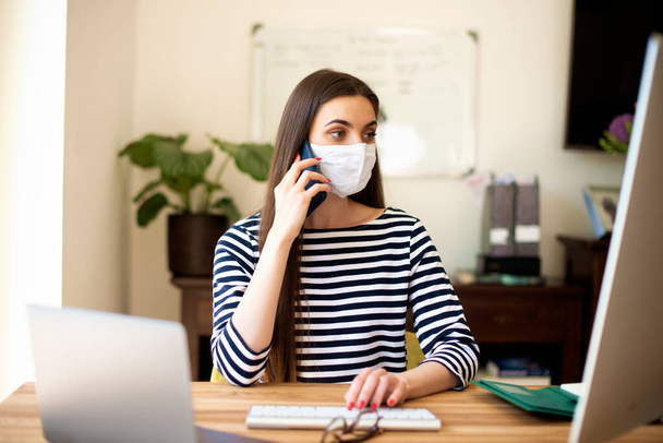 Shot of young businesswoman wearing casual clothes and face mask while working from home. Attractive female sitting at desk and making a call while working on laptop and computer. Home office. - Фото, изображение