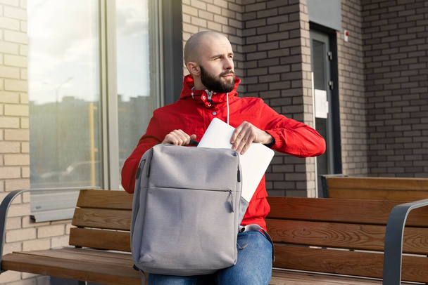 Um cara com barba em roupas casuais tira um computador de sua mochila para trabalhar remotamente. Conceito de trabalhar fora de casa - Foto, Imagem