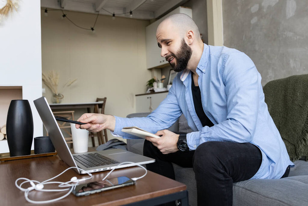 Un joven con una camisa azul y jeans negros estudia online desde casa usando una computadora y un diario para apuntes. Concepto de trabajo y estudio en línea - Foto, Imagen