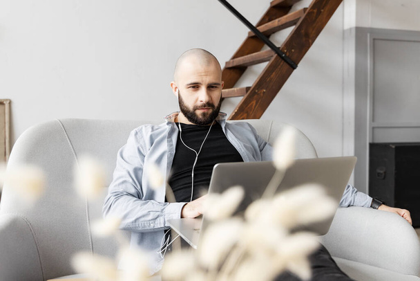 Un tipo con una camisa azul y jeans negros está sentado en el sofá y trabajando en una computadora. El concepto de trabajo freelance - Foto, imagen