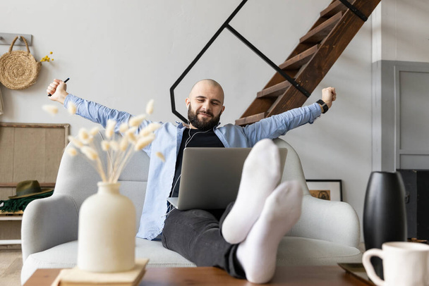 A guy in a blue shirt and black jeans is sitting on the couch and stretching after working at the computer. The concept of freelance work - Фото, изображение