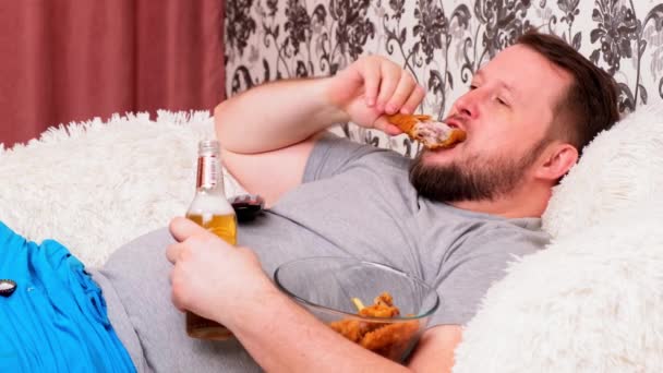 hombre gordo bebe cerveza y come alimentos poco saludables alas de pollo, aburrido delante de la perspectiva de la televisión en el sofá. El concepto de desnutrición, cuarentena en casa, alcoholismo
. - Metraje, vídeo