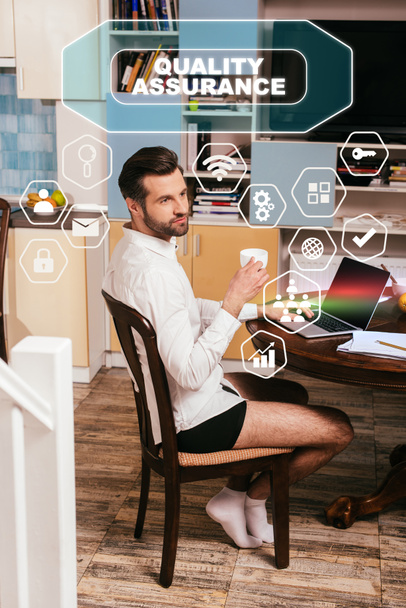Selective focus of handsome man in shirt and panties using laptop and holding cup in living room, quality assurance illustration - Photo, Image