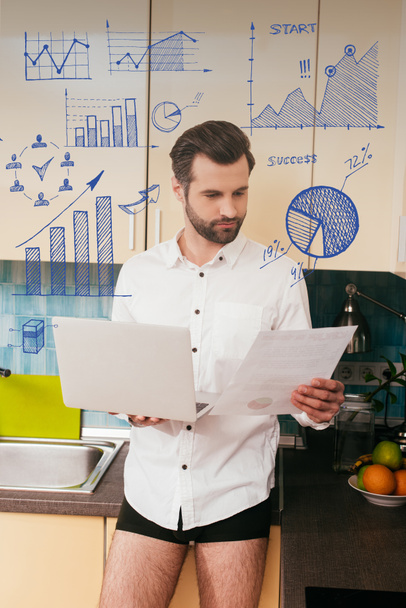 Handsome man in shirt and panties working with document while holding laptop in kitchen, charts and graphs illustration - Valokuva, kuva