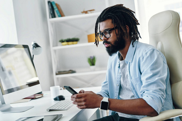 Handsome young African man in eyewear using smartphone while working in the office           - Photo, image