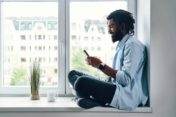 Happy young African man using smartphone and smiling while sitting on the window sill indoors      - Фото, зображення