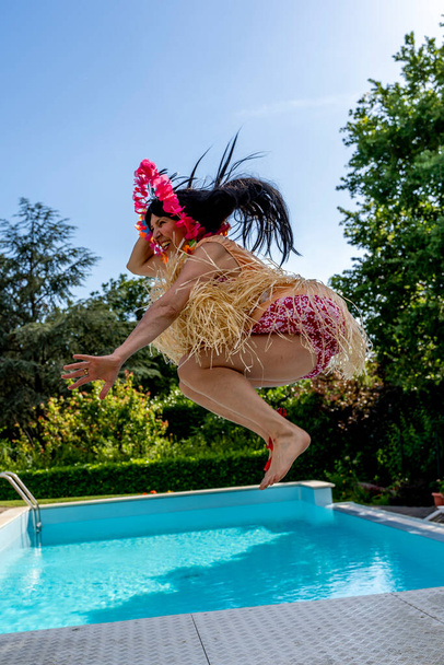 athletic woman dressed in fat Hawaiian costume is jumping upwards by the swimming pool - Foto, afbeelding