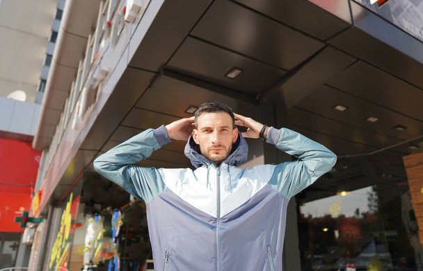 Young attractive guy posing in front of store. Man dressed in three-color suit for training holding hand over head. Concept of modern sports fashion. - Photo, Image
