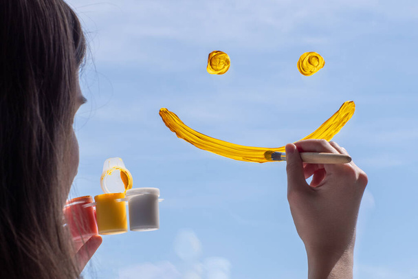 Close-up of a girl draws a yellow smile on glass against a blue sky. The concept of joy, positive, happiness. - Φωτογραφία, εικόνα