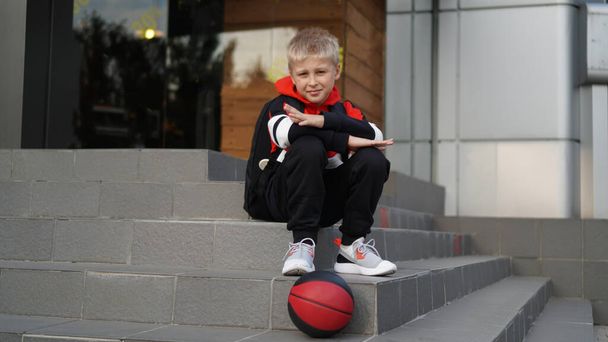 Młody chłopak w kombinezonie treningowym i tenisówkach pozujących z koszykówką przed sklepem. Dziecko siedzi na schodach. Pojęcie młodego sportu i mody. - Zdjęcie, obraz