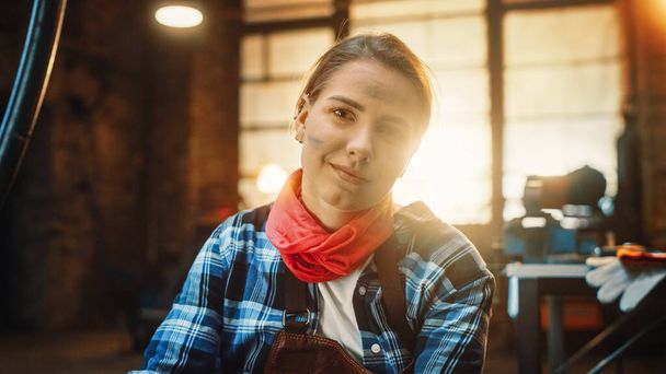 Young Beautiful Empowering Woman with Ear Piercing Gently Smiles at the Camera. Authentic Fabricator Wearing Work Clothes in a Metal Workshop. - Фото, изображение