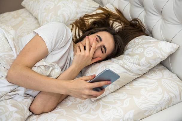 Retrato de mujer joven yacía en la cama con teléfono en el apartamento moderno por la mañana. Despertar o problemas para dormir - Foto, Imagen