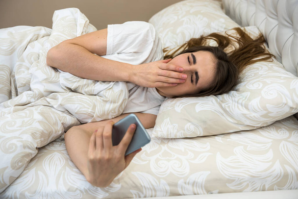 Porträt einer jungen Frau lag am Morgen mit Telefon auf dem Bett in einer modernen Wohnung. Aufwach- oder Schlafprobleme - Foto, Bild