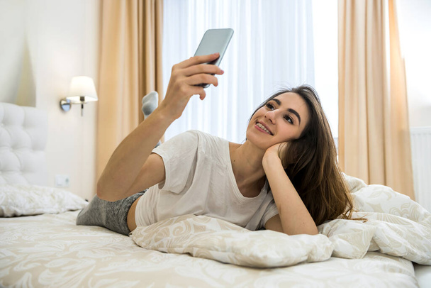 Portret młodej kobiety leżał rano na łóżku z telefonem w nowoczesnym mieszkaniu. Budzenie się lub problemy ze snem - Zdjęcie, obraz
