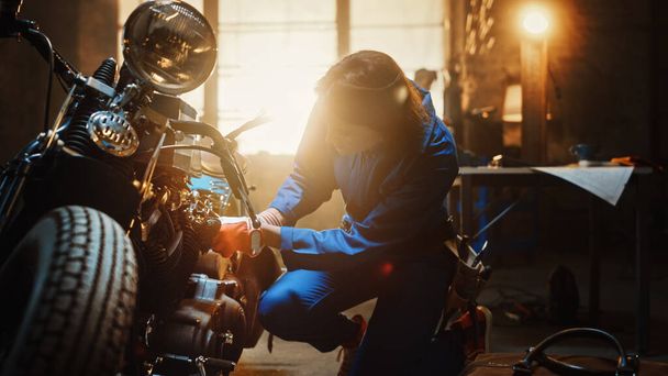 Young Beautiful Female Mechanic is Working on a Custom Bobber Motorcycle. Talented Girl Wearing a Blue Jumpsuit. She Uses a Spanner to Tighten Nut Bolts. Creative Authentic Workshop Garage. - Fotografie, Obrázek