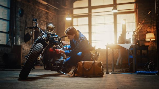 Young Beautiful Female Mechanic is Working on a Custom Bobber Motorcycle. Talented Girl Wearing a Blue Jumpsuit. She Uses a Ratchet Spanner to Tighten Nut Bolts. Creative Authentic Workshop Garage. - Photo, Image