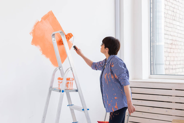 Улыбающаяся женщина средних лет рисует внутреннюю стену дома с помощью краски. Ремонт, реновация, ремонт и освежение квартир. - Фото, изображение