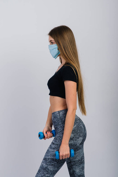 Νεαρή γυναίκα με προστατευτική μάσκα με αλτήρες σε λευκό φόντο. Αθλητικά σε καραντίνα - Φωτογραφία, εικόνα