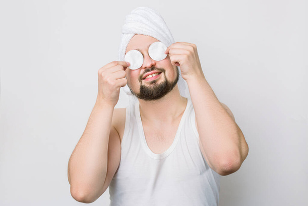 Happy smiling crazy funny bearded man with white towel oh his head, holding two cotton pads and hiding his eyes while caring about his skin. Close up portrait on white background. Spa for men - Photo, Image